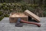 Bearded axe with rosewood handle and leather blade cover resting in front of an open wooden gift box filled with straw