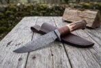 Damascus bowie knife with stag horn and stacked leather handle laid across its brown leather sheath on a rustic table