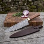 Damascus chef knife resting on an end grain board with red onion and rosemary, brown leather sheath laid on the table in front