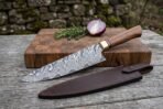 Damascus chef knife with hardwood handle set across a cutting board edge, leather sheath, onion and rosemary behind it outdoors
