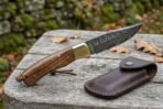 Damascus folding pocket knife with brass bolster resting open on a weathered table next to its snap leather sheath