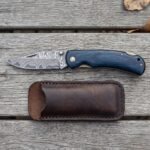 Damascus folding pocket knife with blue wood handle shown open above its brown leather pouch on grey wooden boards