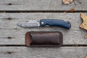 Damascus folding pocket knife with blue wood handle shown open above its brown leather pouch on grey wooden boards