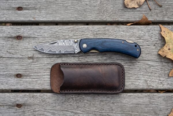 Damascus folding pocket knife with blue wood handle shown open above its brown leather pouch on grey wooden boards