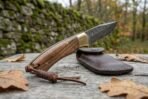 Damascus folding pocket knife with wood handle and brass bolster open on a wooden table, side view with leather pouch
