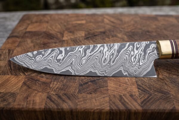 Damascus steel chef knife blade in close view on an end grain cutting board, wavy pattern and brass bolster in focus