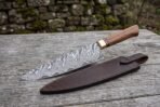 Damascus steel chef knife with hardwood handle and brass bolster lying beside its brown leather sheath on a weathered wooden table