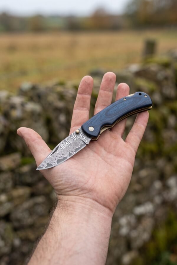 Damascus steel folding knife with blue wood handle held open on an open palm in front of a stone wall and field