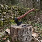 Hand forged bearded axe with rosewood handle stuck in a tree stump with its leather blade cover, by a mossy stone wall