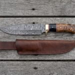 Overhead view of the Damascus bowie with buffalo horn and wood handle lying above its leather sheath on boards