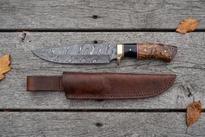 Overhead view of the Damascus bowie with buffalo horn and wood handle lying above its leather sheath on boards