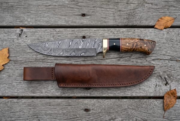 Overhead view of the Damascus bowie with buffalo horn and wood handle lying above its leather sheath on boards