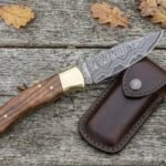 Overhead view of an open Damascus folding knife with wood handle and brass bolster beside a brown leather sheath