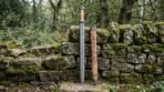 Damascus steel Viking sword standing upright beside its olive burl wooden scabbard against a mossy dry stone wall