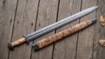 Damascus steel Viking sword and olive burl scabbard with carved steel fittings laid side by side on old wooden boards