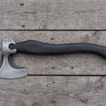 Bearded axe with a brushed steel head and curved black wood handle laid flat on grey weathered planks with wood chips