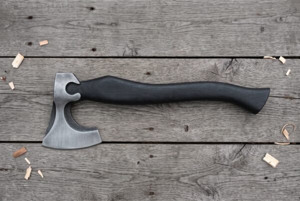 Bearded axe with a brushed steel head and curved black wood handle laid flat on grey weathered planks with wood chips