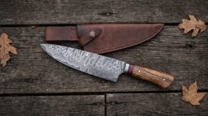 Damascus chef knife with wood handle and red spacer next to its brown leather sheath on dark planks with oak leaves