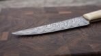 Damascus steel fillet knife with bone handle on a dark end grain cutting board, long thin blade pattern in sharp focus