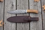 Damascus bowie knife and its long brown leather sheath laid side by side from above on grey planks with leaves