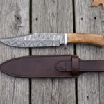 Damascus bowie knife and its long brown leather sheath laid side by side from above on grey planks with leaves