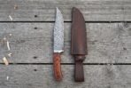 Damascus bowie knife with rosewood handle standing upright next to its dark leather sheath on grey planks, top view