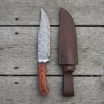 Damascus bowie knife with rosewood handle standing upright next to its dark leather sheath on grey planks, top view