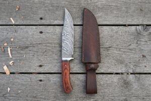 Damascus bowie knife with rosewood handle standing upright next to its dark leather sheath on grey planks, top view