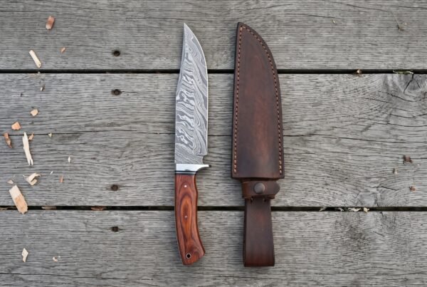 Damascus bowie knife with rosewood handle standing upright next to its dark leather sheath on grey planks, top view