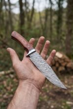 Damascus bowie knife with rosewood handle and steel bolster held in an open hand in woodland, blade pointing down