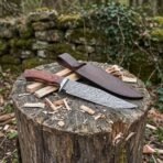 Damascus bowie knife with rosewood handle on a tree stump among wood chips, dark brown leather sheath behind it
