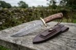 Damascus bowie knife with wood handle, brass guard and pins on a weathered fence rail with its brown leather sheath