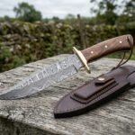Damascus bowie knife with wood handle, brass guard and pins on a weathered fence rail with its brown leather sheath