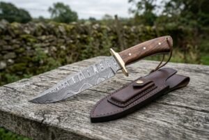 Damascus bowie knife with wood handle, brass guard and pins on a weathered fence rail with its brown leather sheath