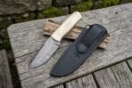 Damascus bull cutter knife with camel bone handle placed beside its black leather sheath on weathered boards