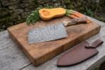 Damascus cleaver with stag horn handle on a butcher block with squash and carrots, brown leather sheath beside it