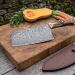 Damascus cleaver with stag horn handle on a butcher block with squash and carrots, brown leather sheath beside it