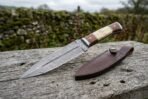 Damascus dagger knife with wood and bone handle propped on a log beside its brown leather sheath outdoors