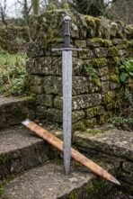 Damascus dagger sword standing upright against a mossy stone wall with its olive burl scabbard leaning on the steps