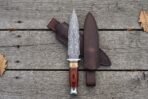 Damascus dagger with wood and bone handle laid flat on grey planks next to its dark brown leather sheath