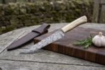 Damascus fillet knife with bone handle on a cutting board with rosemary and garlic, brown leather sheath beside it on the table