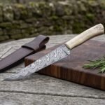 Damascus fillet knife with bone handle on a cutting board with rosemary and garlic, brown leather sheath beside it on the table