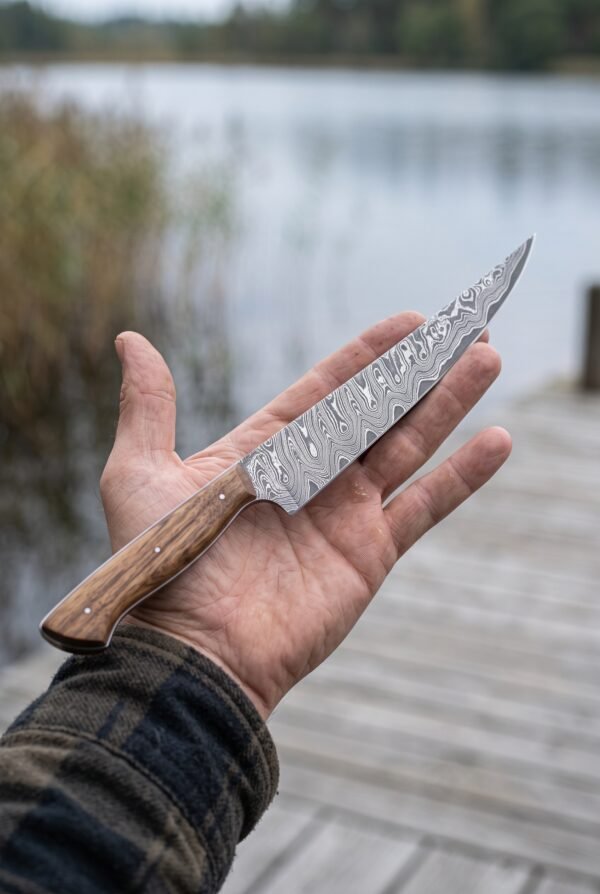 Damascus fillet knife with slim flexible blade and wood handle held in an open hand by a lake