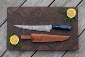 Damascus fillet knife with blue resin handle and tan leather sheath on an end grain board with lemon halves and dill