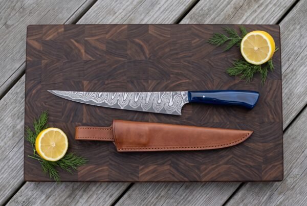 Damascus fillet knife with blue resin handle and tan leather sheath on an end grain board with lemon halves and dill