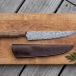 Damascus fillet knife with wood handle on a cutting board with its leather sheath, lemon half and fresh herbs