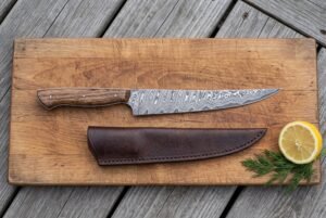 Damascus fillet knife with wood handle on a cutting board with its leather sheath, lemon half and fresh herbs
