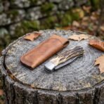 Damascus folding pocket knife with stag horn handle shown closed on a tree stump beside its tan leather sheath