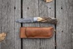 Damascus folding pocket knife with stag horn handle lying open on grey boards from above, leather sheath below it