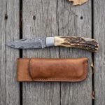 Damascus folding pocket knife with stag horn handle lying open on grey boards from above, leather sheath below it